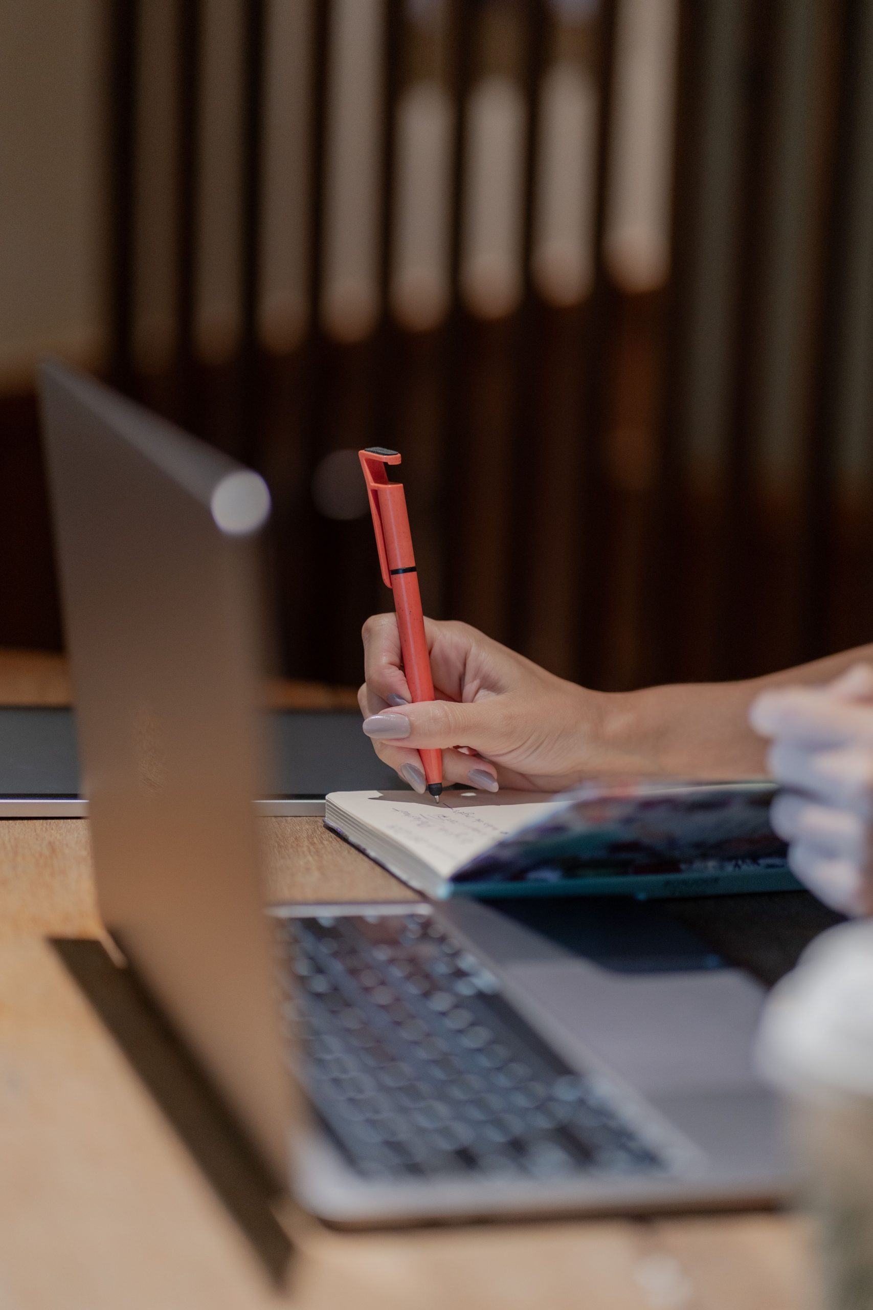 Adult writing in a notebook next to a laptop on a wooden desk indoors.